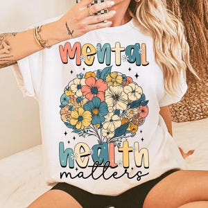 May include: White t-shirt with the words "Mental Health Matters" in a colorful, floral design. The design features a bouquet of flowers in shades of pink, blue, yellow, and white. The shirt has short sleeves and a crew neck.
