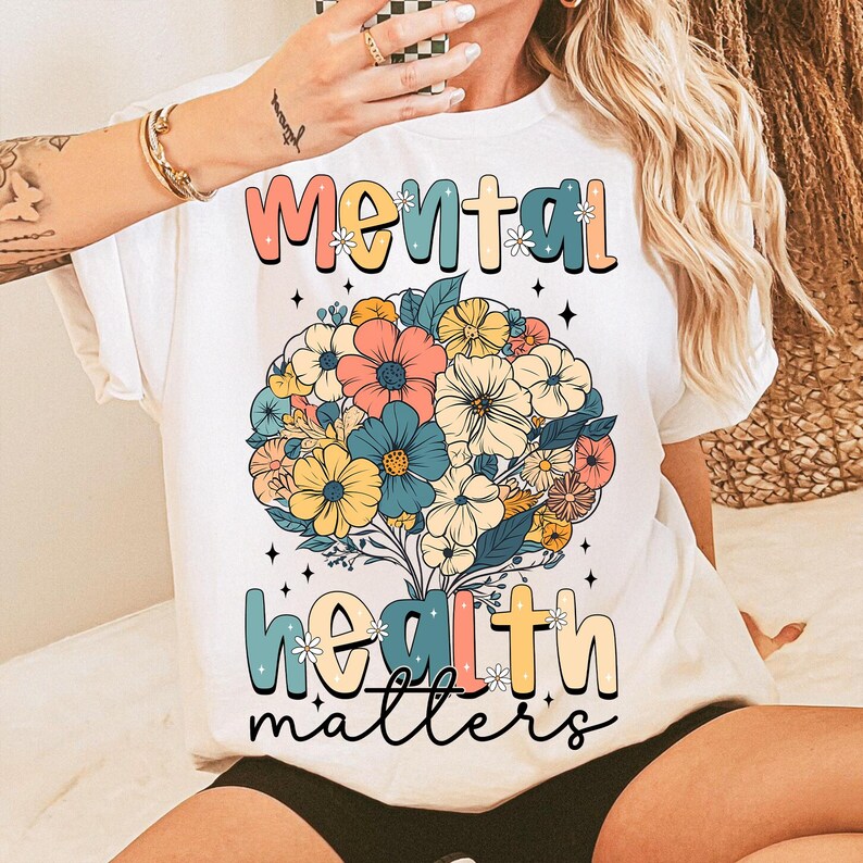 May include: White t-shirt with the words "Mental Health Matters" in a colorful, floral design. The design features a bouquet of flowers in shades of pink, blue, yellow, and white. The shirt has short sleeves and a crew neck.