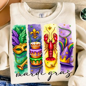 May include: A white sweatshirt with a colorful Mardi Gras design. The design features a purple, green, and gold mask, a cake with purple frosting, a red lobster, and a purple and gold hat. The text "Mardi Gras" is printed below the design.