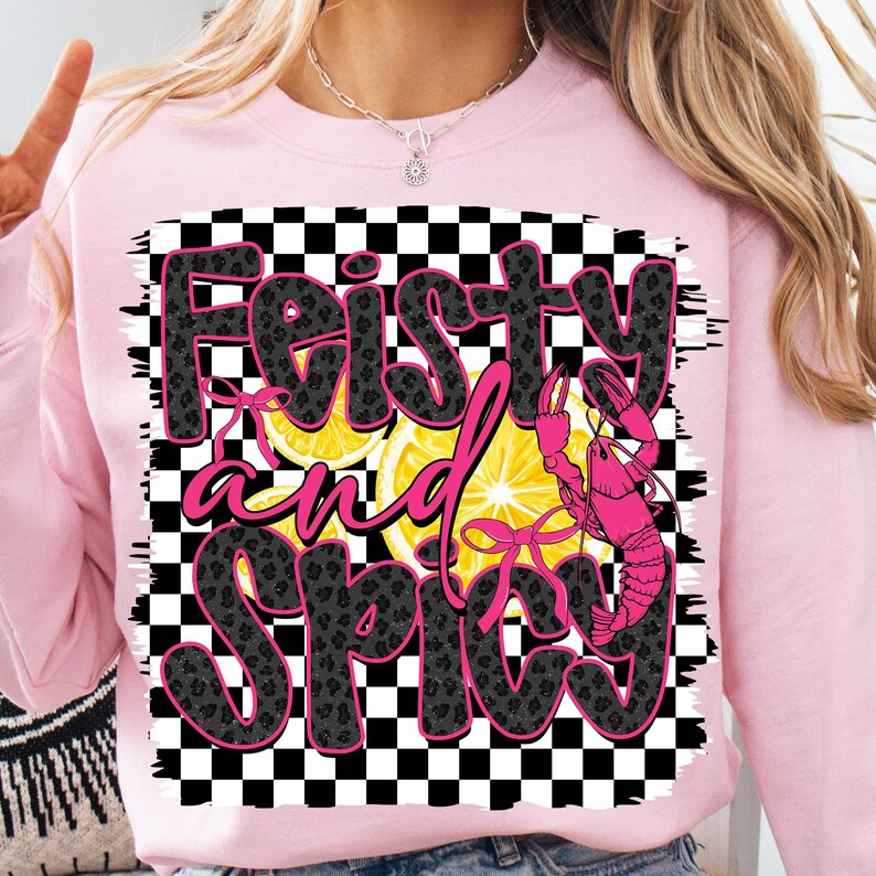 May include: Pink sweatshirt with a black and white checkered background. The text "Feisty and Spicy" is written in pink with a black leopard print pattern. A pink crawfish and two lemon slices are on the checkered background.