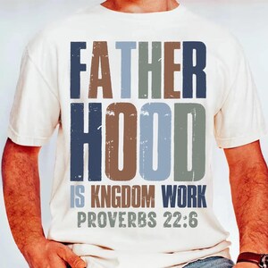 以下が含まれることがあります： クリーム色のTシャツ。テキスト「FATHERHOOD IS KINGDOM WORK PROVERBS 22:6」が、使い古されたマルチカラーフォントでプリントされています。テキストは青、茶色、グレーの色合いです。