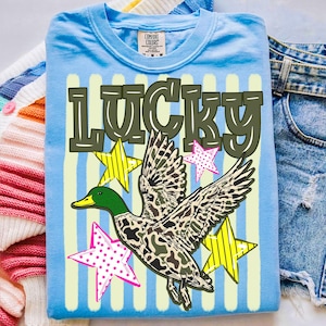 Könnte beinhalten: Ein hellblaues T-Shirt mit dem Wort "LUCKY" in olivgrünen Blockbuchstaben. Eine Ente mit camouflagefarbenen Flügeln und einem grünen Kopf ist abgebildet, zusammen mit gelben und rosa Sternen. Das T-Shirt ist gefaltet.