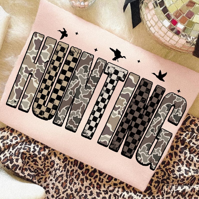 Puede incluir: Una sudadera rosa claro con la palabra "HUNTING" en letras grandes y en negrita. Las letras alternan entre un patr&oacute;n de camuflaje y un patr&oacute;n de cuadros blanco y negro. Peque&ntilde;as siluetas de p&aacute;jaros negros y estrellas est&aacute;n dispersas alrededor de la palabra.
