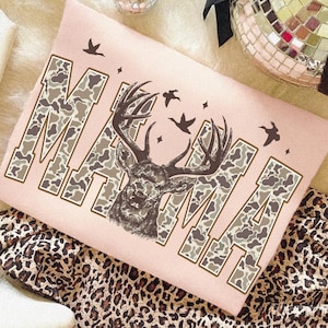 May include: A light pink sweatshirt with the word "MAMA" in a camouflage pattern. A deer head graphic is centered, with birds and stars. The sweatshirt is on a leopard print fabric.