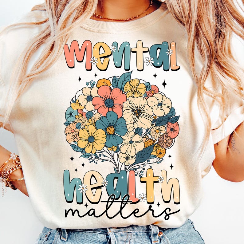 May include: Cream-colored t-shirt featuring the words "Mental Health Matters" in a colorful, retro font. A floral design in shades of pink, blue, yellow, and orange is centered above the words, creating a positive and uplifting message.