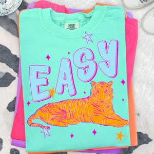 May include: A light teal Comfort Colors t-shirt with the word "EASY" in light blue and a reclining orange tiger graphic. The design includes pink stars and a starburst. The shirt is folded on top of other colorful shirts.