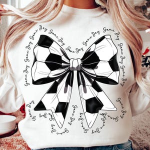 May include: White sweatshirt featuring a large bow design made of soccer balls in black and white. The words "Game Day" are written in a circular pattern around the bow. A sporty and stylish apparel item.