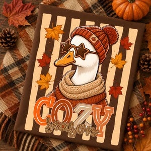 May include: A brown t-shirt featuring a graphic of a white goose wearing an orange knit beanie with a pumpkin icon, star-shaped sunglasses, and a beige scarf. The background has vertical stripes and autumn leaves with the text COZY season.