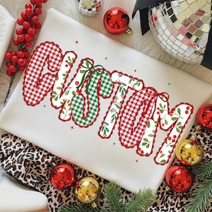 Pu&ograve; includere: Una felpa bianca con la scritta "CUSTOM" in un design festivo. Le lettere sono riempite con motivi a quadri rossi e verdi, motivi floreali e bordi smerlati rossi. Ornamenti natalizi e verde circondano la felpa.