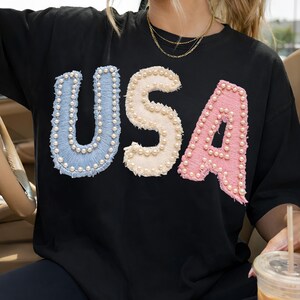 K&ouml;nnte beinhalten: Schwarzes T-Shirt mit dem Wort "USA" in Hellblau, Creme und Rosa, wobei jeder Buchstabe mit kleinen, wei&szlig;en Perlen verziert ist. Das Shirt hat einen Rundhalsausschnitt und kurze &Auml;rmel.
