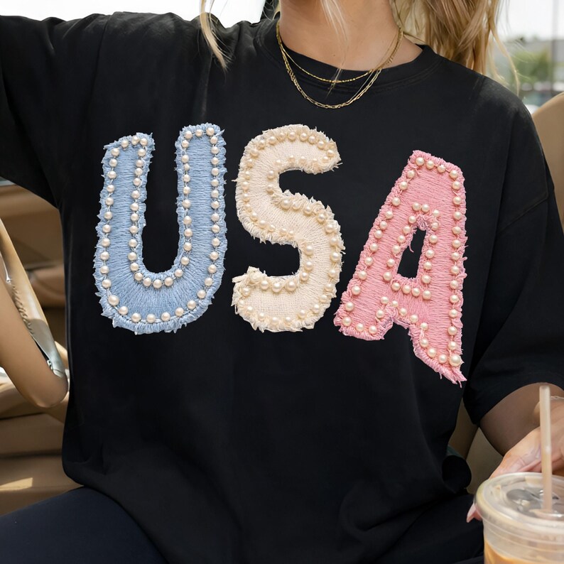 K&ouml;nnte beinhalten: Schwarzes T-Shirt mit dem Wort "USA" in Hellblau, Creme und Rosa, wobei jeder Buchstabe mit kleinen, wei&szlig;en Perlen verziert ist. Das Shirt hat einen Rundhalsausschnitt und kurze &Auml;rmel.