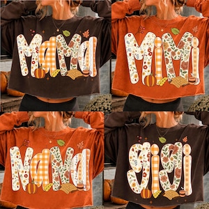 May include: A collection of four long-sleeve crewneck sweatshirts in brown and orange. Each features large block lettering spelling Mama, Mimi, Nana, or Gigi, decorated with floral patterns, orange gingham, pumpkins, leaves, and coffee mugs.