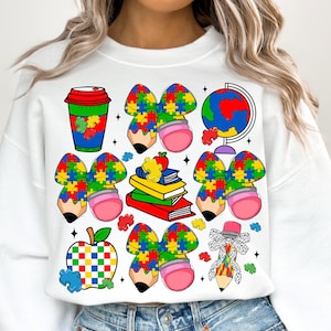 May include: White sweatshirt with a colorful pattern of puzzle pieces, bows, pencils, an apple, a coffee cup, and a globe. The puzzle pieces are in the colors of the autism awareness ribbon.