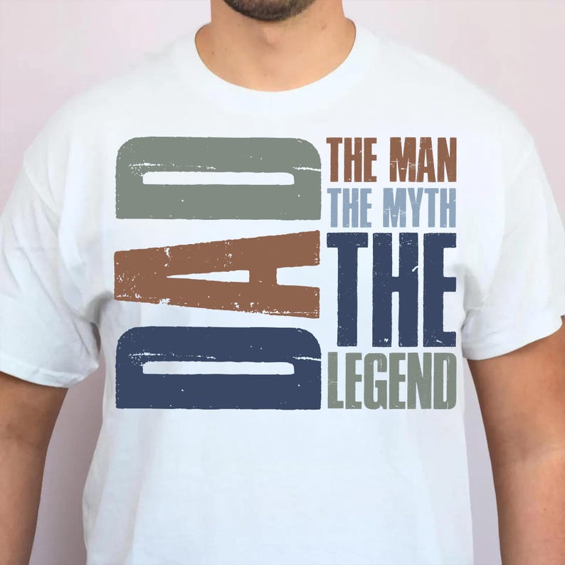 Peut inclure: T-shirt blanc avec le mot "DAD" en grandes lettres us&eacute;es grises, marron et bleues. Le texte "THE MAN THE MYTH THE LEGEND" est imprim&eacute; verticalement dans une palette de couleurs compl&eacute;mentaires.