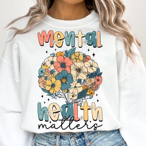 May include: White sweatshirt with a floral design and the text "Mental Health Matters." The floral arrangement features a variety of colorful flowers in shades of peach, blue, yellow, and orange. The text is in a retro font.