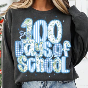 K&ouml;nnte beinhalten: Dunkelgraues Sweatshirt mit einem blau-wei&szlig;en Design mit der Aufschrift "100 Days of School". Die Zahlen und Buchstaben sind mit floralen Mustern und Vichy-Druck verziert. Das Design beinhaltet Schleifen und Sterne.