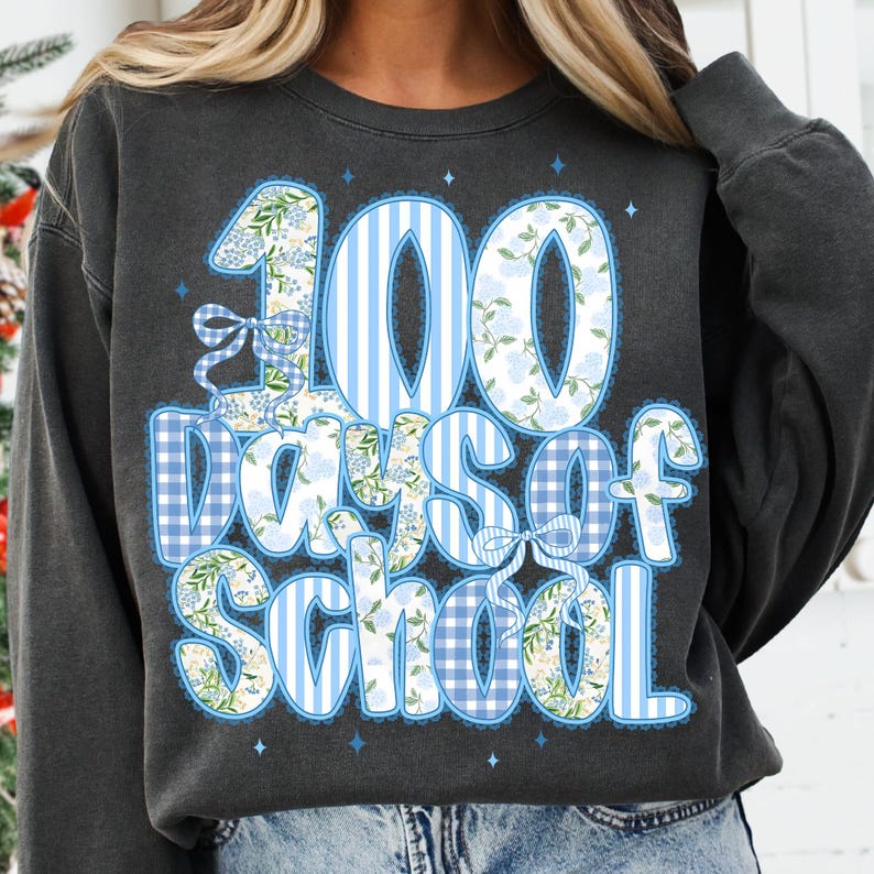 K&ouml;nnte beinhalten: Dunkelgraues Sweatshirt mit einem blau-wei&szlig;en Design mit der Aufschrift "100 Days of School". Die Zahlen und Buchstaben sind mit floralen Mustern und Vichy-Druck verziert. Das Design beinhaltet Schleifen und Sterne.