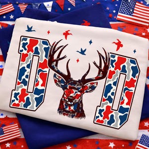 May include: White t-shirt with the word "DAD" in a patriotic red, white, and blue camouflage pattern. A deer head with antlers is centered between the letters, also in the same camouflage design. Blue birds and red diamonds decorate the shirt.