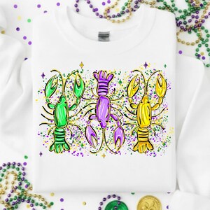 May include: White crew neck sweatshirt featuring three colorful crawfish illustrations. The crawfish are green, purple, and yellow, with black outlines and decorative accents. The design includes colorful paint splatters and star details.