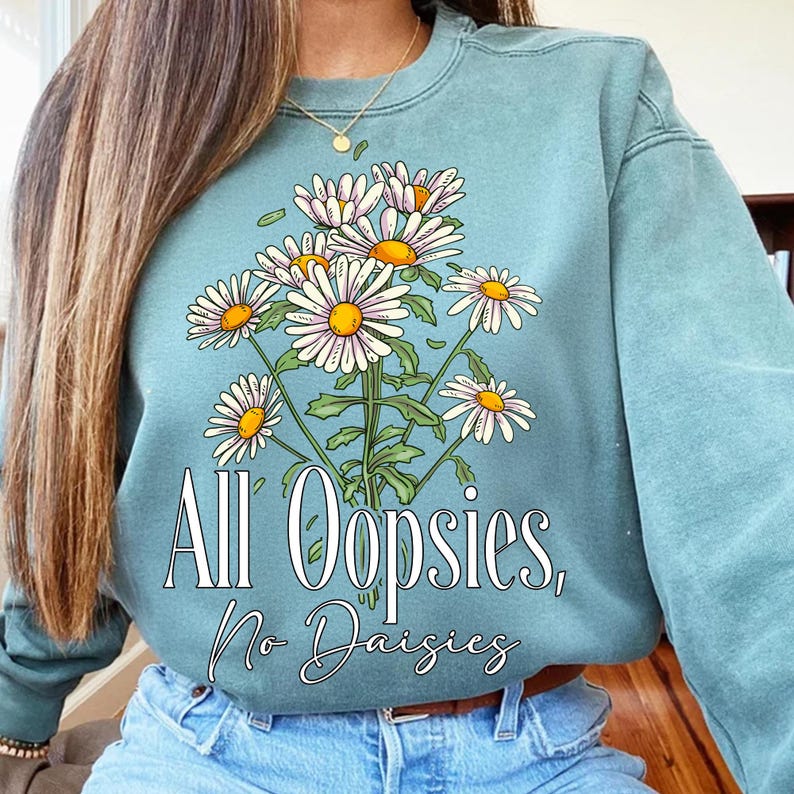 K&ouml;nnte beinhalten: Ein teal-farbenes Sweatshirt mit einem Strau&szlig; G&auml;nsebl&uuml;mchen mit gelben Zentren und wei&szlig;en Bl&uuml;tenbl&auml;ttern. Der Text "All Oopsies, No Daisies" ist unter den Blumen aufgedruckt. Das Sweatshirt hat lange &Auml;rmel.