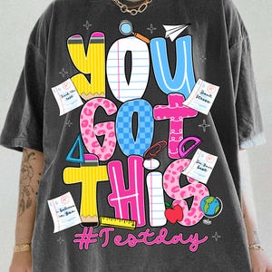 Peut inclure: T-shirt gris foncé avec un motif coloré disant "YOU GOT THIS". Le design comprend des éléments scolaires comme des crayons, des avions en papier et une pomme. Le hashtag "#Testday" est également présent.
