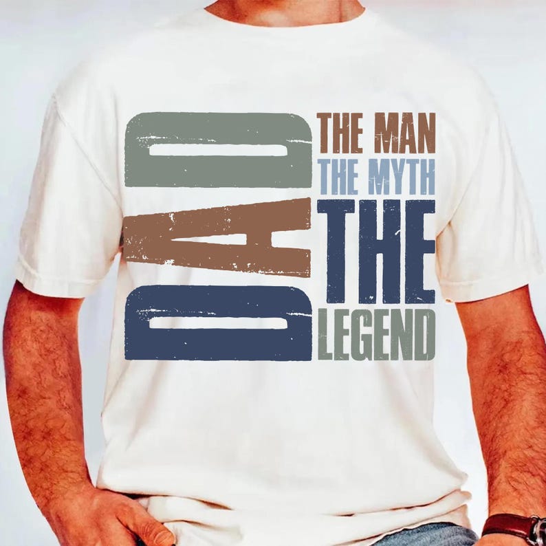 Peut inclure: T-shirt de couleur cr&egrave;me avec le mot "DAD" en grandes lettres majuscules d&eacute;lav&eacute;es. Au-dessus de "DAD" se trouvent les mots "THE MAN THE MYTH THE LEGEND" superpos&eacute;s. Le texte est dans des tons de gris, de marron et de bleu.