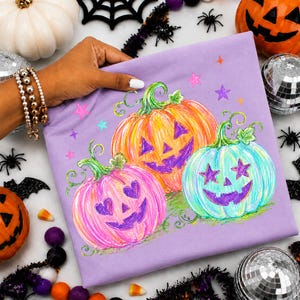 May include: A light purple t-shirt featuring a graphic illustration of three jack-o'-lanterns in pink, orange, and light blue. The pumpkins have carved faces with heart and star-shaped eyes, surrounded by small colorful stars on the fabric.