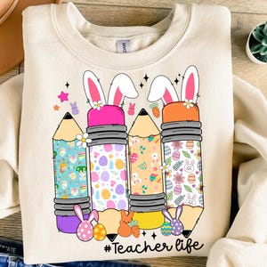 Op de afbeelding: Een beige sweatshirt met een afbeelding van drie potloden met oren van een paas konijn. De potloden zijn versierd met kleurrijke paasei-patronen. De tekst "#Teacher life" is onder de potloden gedrukt.