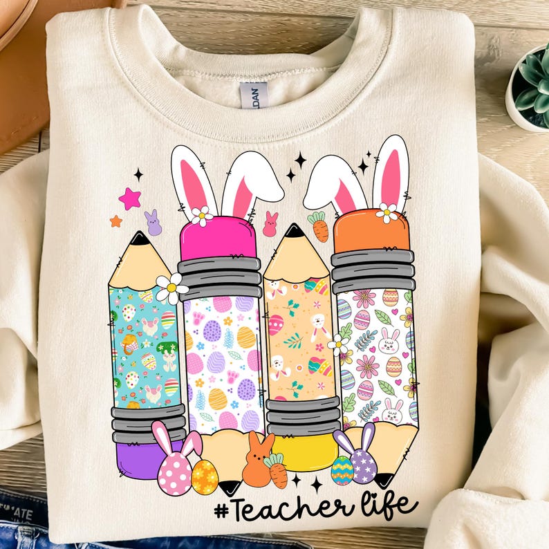 Op de afbeelding: Een beige sweatshirt met een afbeelding van drie potloden met oren van een paas konijn. De potloden zijn versierd met kleurrijke paasei-patronen. De tekst "#Teacher life" is onder de potloden gedrukt.