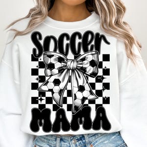 May include: White sweatshirt with the words "Soccer Mama" in black, featuring a soccer ball bow design. The background has a black and white checkered pattern with star accents. A casual and sporty design.
