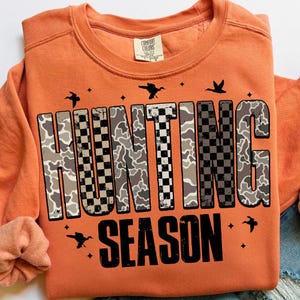 Puede incluir: Sudadera naranja Comfort Colors con las palabras "HUNTING SEASON" en un patr&oacute;n de camuflaje y cuadros. El dise&ntilde;o incluye siluetas de p&aacute;jaros y estrellas. La sudadera tiene mangas largas y cuello redondo.