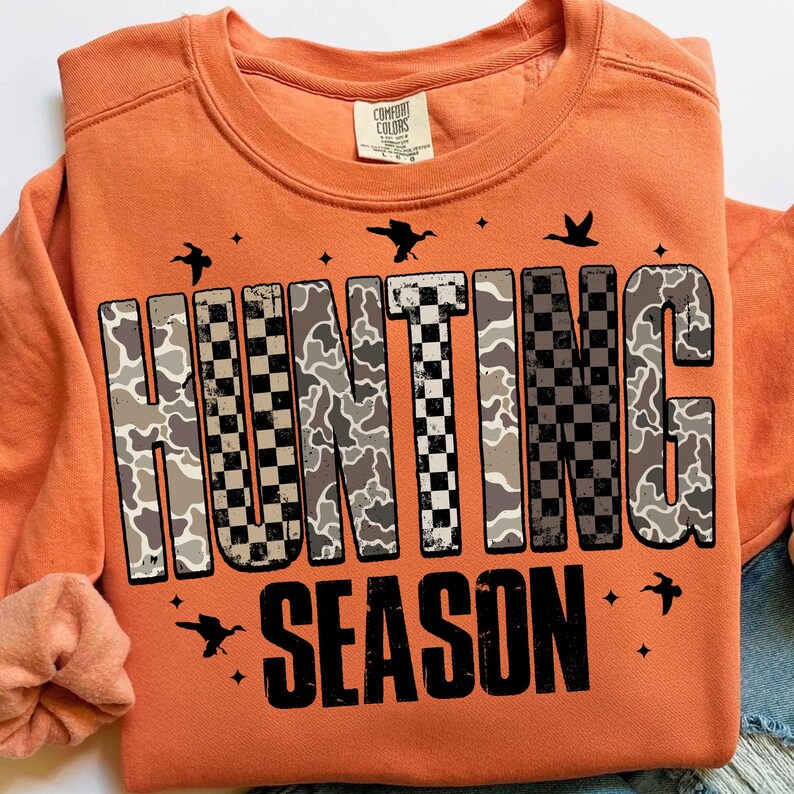 Puede incluir: Sudadera naranja Comfort Colors con las palabras "HUNTING SEASON" en un patr&oacute;n de camuflaje y cuadros. El dise&ntilde;o incluye siluetas de p&aacute;jaros y estrellas. La sudadera tiene mangas largas y cuello redondo.