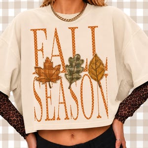 Könnte beinhalten: Ein cremefarbenes Kurzarm-T-Shirt mit dem Schriftzug FALL SEASON in großen, orangefarbenen Buchstaben mit Punkten. Drei stilisierte Blätter mit orangefarbenen, grünen und gelben Karomustern liegen über dem Text. Getragen über einem langärmeligen Leoparden-Oberteil.