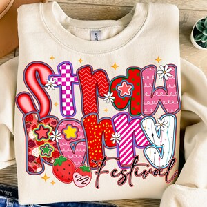May include: A cream-coloured sweatshirt with the words "Strawberry Mommy Festival" in a colourful graphic. The text incorporates strawberry, star, and daisy patterns. The design is outlined in red and black.