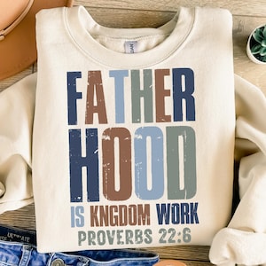 以下が含まれることがあります： クリーム色のスウェットシャツ。"FATHER HOOD IS KINGDOM WORK PROVERBS 22:6"の文字がかすれたフォントでプリントされています。テキストは青、茶色、緑の色合いです。スウェットシャツはクルーネックです。