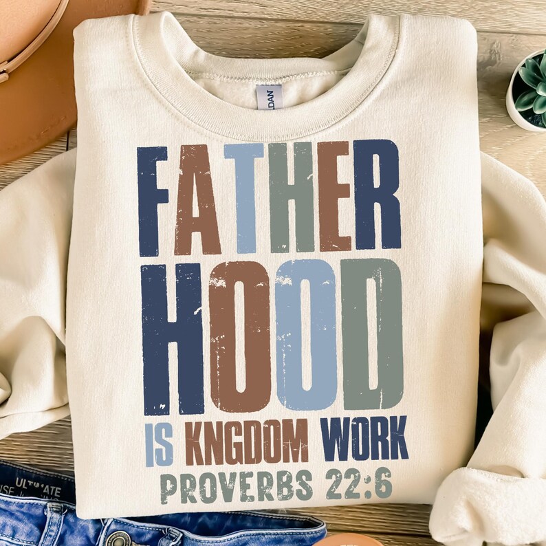 以下が含まれることがあります： クリーム色のスウェットシャツ。"FATHER HOOD IS KINGDOM WORK PROVERBS 22:6"の文字がかすれたフォントでプリントされています。テキストは青、茶色、緑の色合いです。スウェットシャツはクルーネックです。