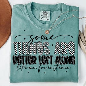 Puede incluir: Una camiseta Comfort Colors turquesa con el texto "Some Things Are Better Left Alone, like me, for instance." La palabra "THINGS" está en un patrón de cuadros blancos y negros. Un collar plateado con un colgante está sobre la camiseta.