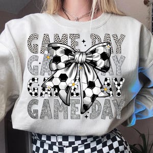 May include: A light gray sweatshirt with the words "GAME DAY" in various fonts. A large bow with a soccer ball pattern is in the center, adorned with small yellow flowers. The sweatshirt is a casual style.
