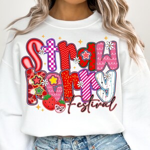 May include: White sweatshirt featuring the text "Strawberry Festival" in a colourful, playful font. The letters are decorated with strawberries, stars, and floral accents. The design is vibrant and eye-catching.