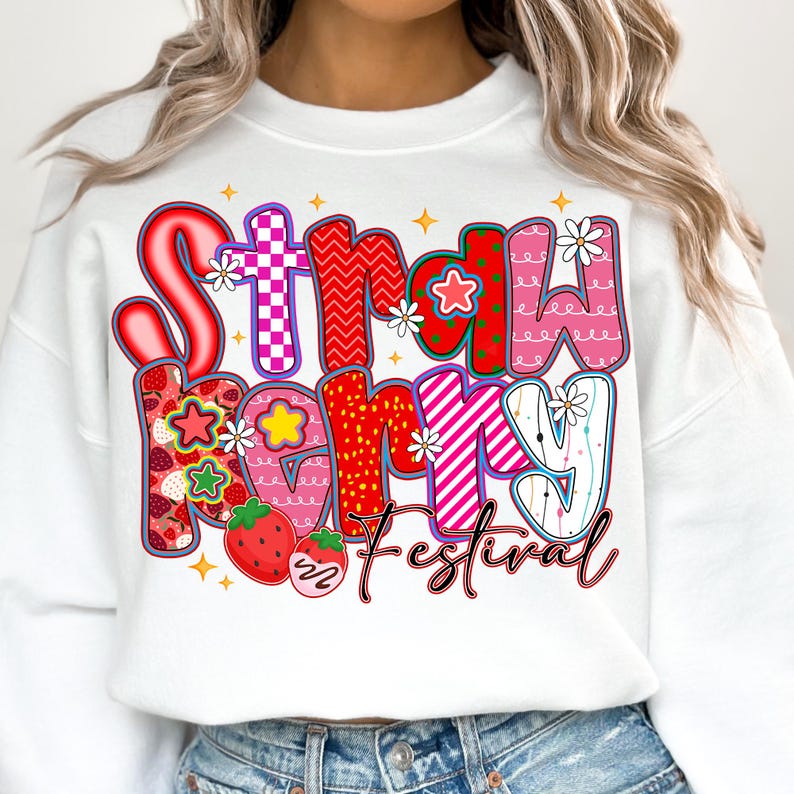 May include: White sweatshirt featuring the text "Strawberry Festival" in a colourful, playful font. The letters are decorated with strawberries, stars, and floral accents. The design is vibrant and eye-catching.
