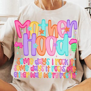 May include: A white t-shirt with the word "Motherhood" in colorful bubble letters, decorated with flowers. Below, the text reads "Some days I rock it, some days it rocks me, either way we're rockin".