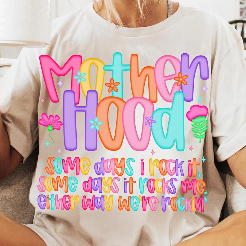 May include: A white t-shirt with the word "Motherhood" in colorful bubble letters, decorated with flowers. Below, the text reads "Some days I rock it, some days it rocks me, either way we're rockin".
