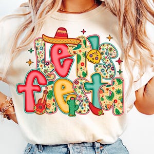 May include: Cream-colored t-shirt with the words "Let's Fiesta" in a colorful, festive design. The design includes a sombrero, guitar, cactus, skull, and chili pepper, all with floral patterns.