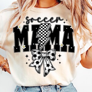 May include: Cream-colored t-shirt with the words "soccer MAMA" in black block letters. A checkered lightning bolt graphic is centered between the words. A soccer ball bow is below the text, with small white flowers and stars.