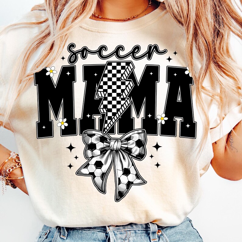 May include: Cream-colored t-shirt with the words "soccer MAMA" in black block letters. A checkered lightning bolt graphic is centered between the words. A soccer ball bow is below the text, with small white flowers and stars.