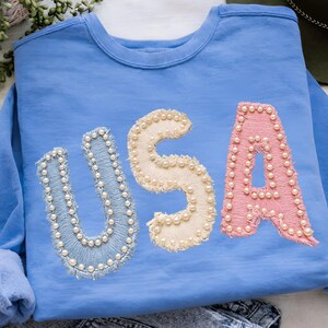K&ouml;nnte beinhalten: Hellblaues Sweatshirt mit dem Wort "USA" auf der Vorderseite. Die Buchstaben sind in einer Mischung aus Farben gehalten: Hellblau, Creme und Rosa, und mit Kunstperlen verziert. Das Sweatshirt hat einen Rundhalsausschnitt und lange &Auml;rmel.