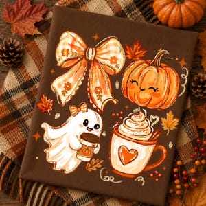 May include: A dark brown t-shirt featuring a graphic print of a large orange gingham bow, a smiling pumpkin, a white ghost holding a coffee cup, and a mug of whipped cream with a heart design, surrounded by small orange maple leaves and stars.