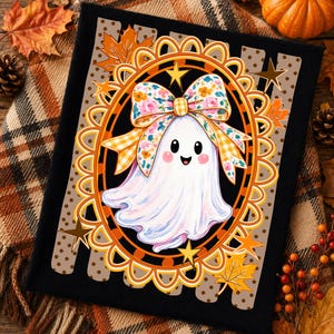 May include: A black t-shirt featuring a graphic of a white ghost with a smiling face, wearing a large floral and yellow gingham patterned bow. The ghost is centered within an ornate orange oval frame decorated with small yellow stars and maple leaves.