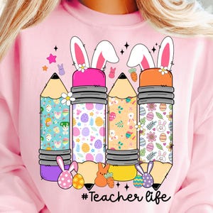 Op de afbeelding: Roze sweatshirt met een afbeelding van drie potloden met konijnenoren. Elk potlood is versierd met een paasei-patroon. De tekst "#Teacher life" is onder de potloden gedrukt.