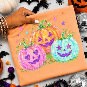 May include: An orange t-shirt featuring a graphic illustration of three jack-o'-lanterns in pink, orange, and light blue. The pumpkins have purple carved faces with heart and star-shaped eyes, surrounded by small purple, pink, and blue stars.